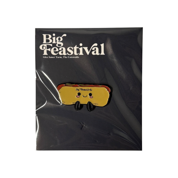 A Hotdog Pin Badge by 2025, showing a cartoon hotdog with a smiling face and tiny legs, attached to a card that says Big Feastival on top against a dark background.