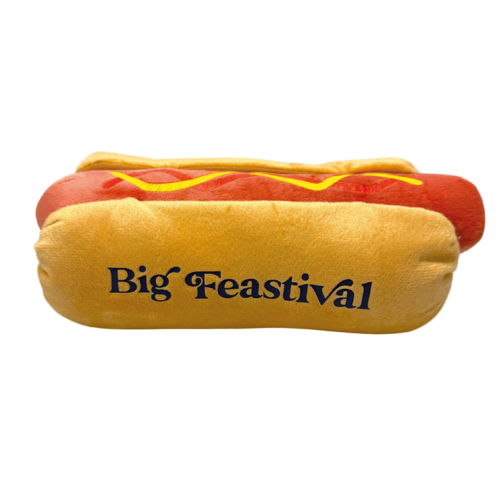 The 2025 Hotdog Plush is a toy shaped like a hot dog with mustard and “Big Feastival” on the bun, shown on a plain white background.